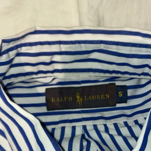 Ralph Lauren Skjorta - Snygg skjorta från Ralph Lauren i storlek S med klassiskt blått och vitt randigt mönster. Skjortan har långa ärmar, knappar framtill och den ikoniska broderade loggan på bröstet. Tillverkad i mjuk bomull som känns skön mot huden.
