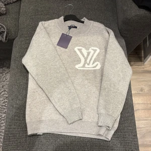 Grå stickad tröja Louis Vuitton M - Supersnygg grå stickad tröja från Louis Vuitton i storlek M. Tröjan har ett stort vitt LV-logo broderat både fram och bak, ribbad rund hals och muddar vid ärmslut och nederkant. Perfekt för dig som gillar exklusiva märken och stilren design.