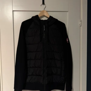 Svart cardigan från Canada Goose - Svart cardigan från Canada Goose med huva och snörning. Jackan har quiltad framsida, dragkedja och klassisk logotyp på ärmen. Materialet är polyester och jackan har en clean, modern look som funkar till streetwear. Perfekt för kyliga dagar. Nypris ligger mellan 7500-8000.