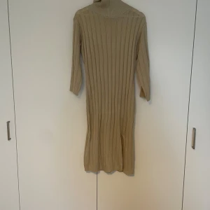 Beige ribbad stickad långklänning - Säljer en beige stickad långklänning med polokrage och ribbad struktur. Klänningen har trekvartsärm och en liten slits framtill. Perfekt för dig som gillar en enkel men snygg look med lite extra edge.
