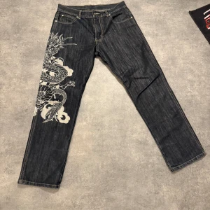 Mukarachu jeans med drakmotiv, stl 30 - Unika svarta jeans från Mukarachu i storlek 30 med stort broderat och tryckt drakmotiv i vitt, gult och orange längs benet. Klassisk femficksmodell med raka ben och snygga kontrastsömmar. Perfekta för dig som vill sticka ut med coola detaljer.