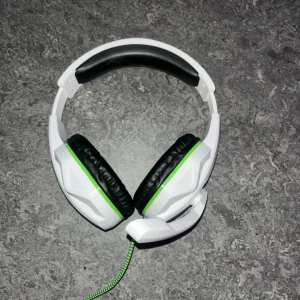 Kabelanslutet gaming headset med mikrofon - Snyggt gaming headset i vitt och grönt med over-ear design och inbyggd mikrofon. Kabelanslutning med 3,5 mm kontakt och adapter för PC. Mjuka öronkuddar och justerbar bygel. Ser ut att vara i gott skick utan synligt slitage.