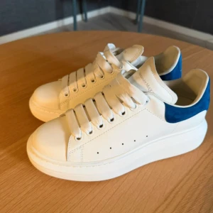 Alexander McQueen vita sneakers med blå mocka - Säljer ett par snygga Alexander McQueen sneakers i vitt skinn med blå mockadetalj på hälen. Skorna har breda vita snören, platt sula med coolt mönster undertill och rund tå. Perfekta för dig som gillar stilrena och trendiga sneakers med lyxig känsla. Hör av er vid minsta frågor eller funderingar. Box medgår inte pågrund av att den gått sönder så jag slängde den