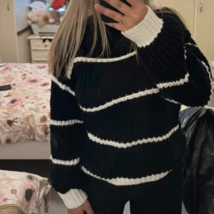 Svart stickad tröja med vita ränder - Mysig svart stickad tröja med vita horisontella ränder och kontrasterande vita muddar. Tröjan har en loose passform och långa ärmar, perfekt för kyligare dagar. Enkel att matcha med jeans eller kjol.
