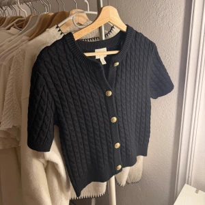 Svart kabelstickad kofta från H&M - Svart kabelstickad kofta från H&M med korta ärmar och guldfärgade knappar. Köpt utomlands och helt oanvänd 💗