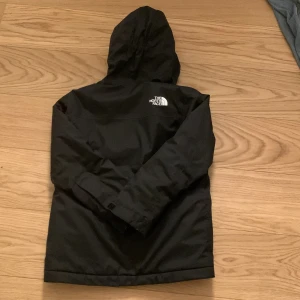 Svart vindjacka från The North Face - Svart vindjacka från The North Face med huva och vit logga på bröstet. Jackan har dragkedja framtill och är tillverkad i slitstark polyester. Perfekt för blåsiga dagar och har en clean, sportig look med långärmade ärmar och normal passform.