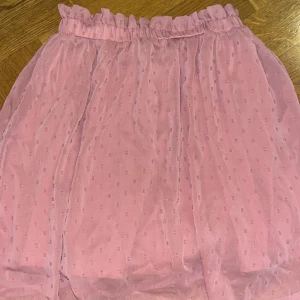 Rosa prickig kortkjol från H&M Divided - Söt rosa kortkjol från H&M Divided med små prickar i tyget och volangkant upptill. Kjolen har en elastisk midja och är gjord i ett lätt, transparent polyestermaterial med underkjol. Perfekt för dig som gillar romantiska och lekfulla detaljer.