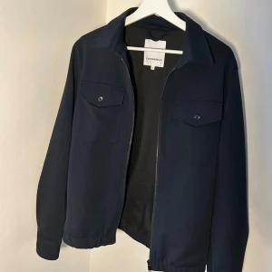 Mörkblå overshirt från Lindbergh - Snygg mörkblå overshirt från Lindbergh med klassisk krage, två bröstfickor med knapp och dragkedja framtill. Jackan har långärmad passform och är tillverkad i ett mjukt, slätt material som ger en clean look. Perfekt för dig som gillar stilrena plagg.