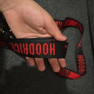 Svart och rött HOODRICH bälte med väska - Säljer ett svart bälte från HOODRICH med röd logotyp längs hela bandet. Bältet har en robust metallspänne och en tillhörande svart väska med vit och röd HOODRICH-tryck. Perfekt accessoar för streetwear-stilen.