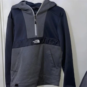 Grå och mörkblå hoodie från The North Face - Säljer en snygg hoodie från The North Face i grått och mörkblått med halv dragkedja och huva. Tröjan har logga på bröstet och ryggen, praktiska fickor och långärmade ärmar. Perfekt för chill eller träning, riktigt skön och stilren design.