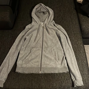 Grå hoodie med dragkedja och huva - Säljer en enkel grå hoodie med dragkedja framtill och huva med snörning. Tröjan har långa ärmar, ribbade muddar och två fickor på framsidan. Perfekt för chill dagar eller när du vill ha något bekvämt och stilrent.