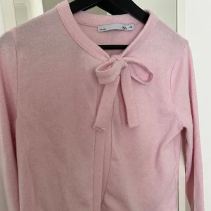 Ljusrosa kofta med rosett från 157 - Supersöt ljusrosa kofta från 157 i storlek XS. Koftan har en fin rosett vid halsen och är gjord i mjukt stickat material. Långärmad och perfekt för dig som gillar gulliga detaljer och en feminin look. Passar dig som vill ha något enkelt men ändå extra.