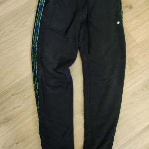 Svarta mjukisbyxor från Champion - Svarta mjukisbyxor från Champion med grönt och blått band längs sidorna där Champion-loggan syns. Byxorna har dragsko i midjan och liten broderad logga på låret. Perfekta för chill eller träning.