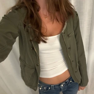 Grön jacka från Abercrombie & Fitch - En olivgrön jacka från Abercrombie & Fitch med dragkedja, knappar och flera fickor framtill. Jackan har rund halsringning och axeldetaljer. Storlek S men något mindre i storleken. 