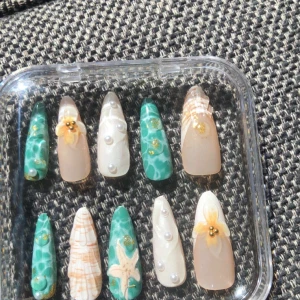 Somriga lösnaglar med pärlor och blommor - Säljer ett set med 10 lösnaglar i tropisk stil. Naglarna har turkosa och beige marmorering, dekorerade med små pärlor, guldfärgade detaljer, 3D-blommor och en sjöstjärna. Perfekt för dig som vill ha unika och kreativa naglar. Kommer i en genomskinlig ask.