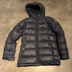 Mörkblå Peak Performance dunjacka - En dunjacka från peak performance. Den har hål på visa ställen men som vi har lagat men tejp. Har lite små hål som syns på bilden. Ena dragkedjan till fickan är sönder och dragkedjan fram kan vara svår att stänga men går att stänga, annars funkar den bra. Säljer billigt för vill få iväg den. 