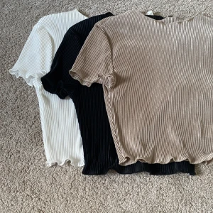 Tre ribbade toppar i olika färger - Säljer tre ribbade toppar med kort ärm i färgerna beige, svart och vit. Alla har en tight passform och är tillverkade i ett glansigt, stretchigt material med vågig kant vid ärmslut och nederkant. Perfekta att styla med jeans eller kjol för en trendig look.