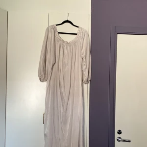 Ljusbeige långklänning från H&M - Säljer en ljusbeige långklänning från H&M i storlek M (oversized). Klänningen har en rundad, rynkad halsringning med snörning framtill och trekvartsärmar med resår. Materialet är mjukt och har en lätt glansig finish som ger en elegant vibe.