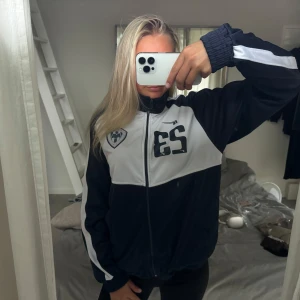 Blå och vit jumper Millennium - Sportig jumper från Millennium i blå och vitt med dragkedja framtill, hög krage och tryckta detaljer på bröstet. Jackan har vita ränder längs ärmarna och en relaxed passform. Perfekt för dig som gillar en clean och stilren look.☺️ Storlek s/m men passar mig med xs.