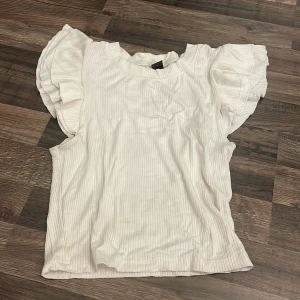 Vit ribbad topp med volangärm från SHEIN - Supersöt vit topp från SHEIN i storlek S, med tunna vertikala ränder och breda volangärmar som ger en extra touch. Materialet är ribbat, perfekt för dig som gillar en chill och feminin stil. Rund halsringning och loose passform.