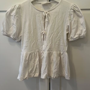 Vit blus med knyt från Gina Tricot - Supersöt vit blus från Gina Tricot med puffiga korta ärmar och volang nedtill. Framsidan har tre knytband som ger en gullig och trendig look. Tillverkad i mjuk bomull.