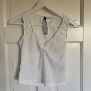 Vit linnetopp med rosett från H&M - Superfin vit linnetopp från H&M Divided med v-ringning och liten söt rosett framtill. Toppen är ärmlös och har en lätt strukturerad, krinklad yta som ger en cool känsla. Perfekt att styla med jeans eller kjol för en fräsch look.