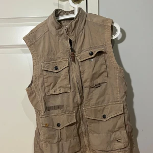 Beige utilityväst  - Säljer en beige utilityväst från Carhartt med flera praktiska fickor framtill och hög krage. Västen har dragkedja och tryckknappar, perfekt för dig som gillar funktionell streetwear. Materialet är slitstark bomull och designen är loose fit.