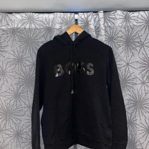 Svart hoodie från BOSS med tryck - Svart hoodie från BOSS med stor, glansig BOSS-logga på bröstet. Tröjan har huva med snörning och långa ärmar. Materialet känns mjukt och bekvämt, perfekt för chill dagar. Klassisk passform och stilren look som funkar till jeans eller joggers.