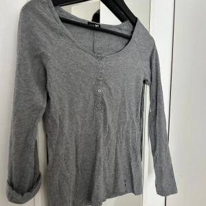 Grå långärmad tröja från JC - Superskön grå långärmad tröja från JC i storlek XS! Tröjan har rund halsringning och flera snygga knappar framtill. Tillverkad i mjuk bomull, perfekt för chill dagar. Ärmarna kan rullas upp och fästas med knapp för en mer avslappnad look.🩶