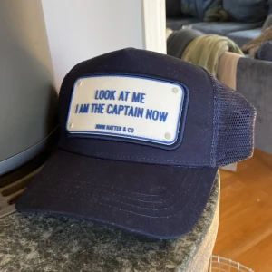 Blå keps John Hatter & Co - Snygg blå keps från John Hatter & Co med mesh på sidorna och bak. Framtill sitter en vit skylt med blå text: 'LOOK AT ME I AM THE CAPTAIN NOW'. Justerbar passform och klassisk trucker-stil. Perfekt statement-piece för dig som vill sticka ut.