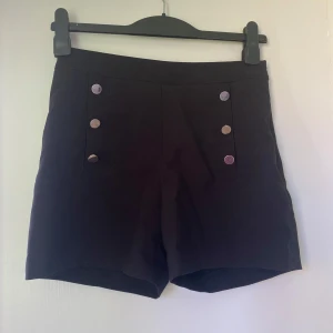 Svarta högmidjade shorts med knappar - Stilrena svarta shorts med hög midja och dekorativa silvriga knappar framtill i dubbla rader. Klassisk och lite marin-inspirerad look. Shortsens material känns slätt och de har diskreta fickor bak samt dragkedja i sidan för en clean finish.