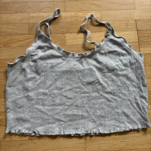 Grå ribbad linnetopp med spetskant - Säljer en ljusgrå croppad linnetopp från Only med tunna axelband och söt spetskant. Toppen är ribbad med små hålmönster och har vågig kant nertill. Perfekt för varma dagar eller att styla lager på lager.