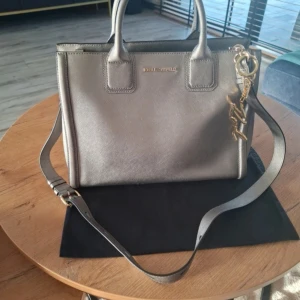 Silver axelväska från Karl Lagerfeld - Snygg axelväska i metallic silver från Karl Lagerfeld med stilren, boxig form och justerbar axelrem. Väskan har två handtag, guldfärgade metalldetMycket gott skick. Använd fåtal gånger av den jag köpte från. Men passar inte min stil. . Väskan har några små fläckar på undersidan men syns inte när man bär. Mått 34×25× 