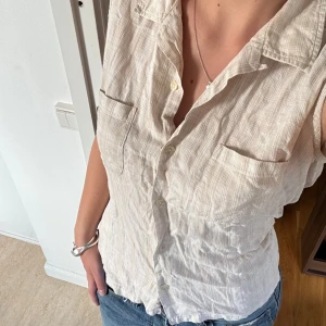 Beige ärmlös skjorta från Columbia - Snygg ärmlös skjorta från Columbia i ljusbeige med diskret randigt mönster. Skjortan har klassisk krage, två bröstfickor och knäppning framtill. Perfekt för varma dagar och enkel att matcha med jeans eller shorts.