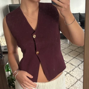 Vinröd stickad väst från Zara - Säljer en vinröd stickad väst från Zara med v-ringning och två snygga guldfärgade knappar framtill. Västen har en asymmetrisk omlott-design och är ärmlös, perfekt att styla över en skjorta eller t-shirt.