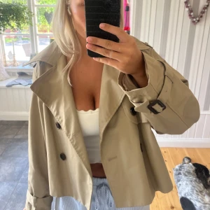 Kort beige trenchcoat - Säljer en snygg croppad trenchcoat i beige med stora slag, dubbelknäppning och bälte vid ärmsluten. Jackan har en loose passform och är perfekt för vår och höst. Materialet känns som polyester eller bomull, riktigt klassisk vibe.