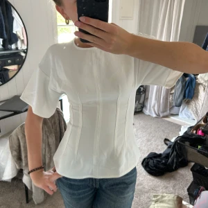 Figursydd t shirt  - Säljer en vit figursydd topp med. Toppen har markerade sömmar som ger en snygg formad siluett och stängs med guldig dragkedja bak. Perfekt för dig som gillar stilrena och trendiga plagg. Använd 1 gång. 