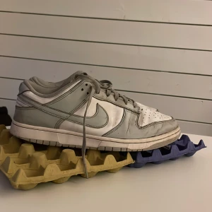 Nike Dunk Low i vitt och grått Helt okej skick Retail pris 1495kr - Nike Dunk Low sneakers i vitt och ljusgrått skinn med klassisk Swoosh på sidorna. Lågt skaft, perforerad tå och snörning framtill. Snygg och tidlös modell som passar till det mesta. Perfekt för dig som gillar streetwear och vill ha en clean look.