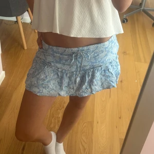 Blå kortkjol med snörning och volang - Supersöt ljusblå kortkjol med vitt paisleymönster, snörning i midjan och volangkant. Det är ”inbyggda” shorts i kjolen, så den är super bekväm och skön!!💕💕