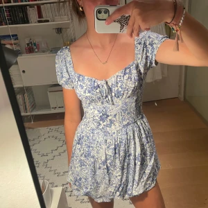 Blommig blåvit kortklänning - Supersöt kortklänning med blått och vitt blommigt mönster, puffärmar och markerad midja. Klänningen har en fyrkantig ringning och öppen rygg. Materialet känns lätt och somrigt, perfekt för varma dagar. Passformen är normal och kjolen är voluminös. Shorts i klänningen.