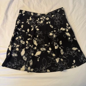 Svart blommig kortkjol med knyt - Supersnygg svart kortkjol med vita blommor och knyt i midjan. Kjolen har en lätt utsvängd form som ger en skön och avslappnad vibe. Perfekt för dig som gillar en feminin och trendig look.