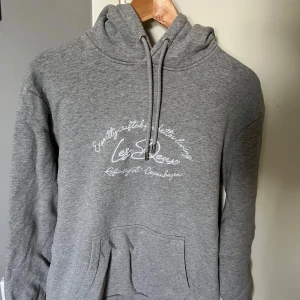 Grå hoodie Les Deux med tryck - Snygg grå hoodie som är slim fit från Les Deux med vit broderad text på bröstet. Tröjan har huva med snörning och en klassisk känguruficka framtill. Perfekt för en chill och avslappnad stil. Köpte för 1599 och säljer den för 500kr och den är helt sprillans ny