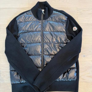 Svart cardigan jacka från Moncler L - Svart cardigan jacka från Moncler med quiltad, glansig front och stickade ärmar och sidor. Jackan har dragkedja framtill och Moncler-logga på ärmen. Perfekt för dig som vill ha en stilren och exklusiv look med sportig touch.