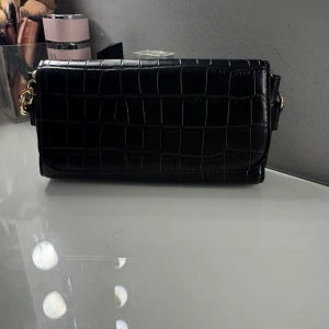 Svart axelväska med kedja - Snygg svart axelväska i croco-mönstrat skinnimitation med guldig kedjerem. Väskan har en rektangulär form och stängs med lock. Går även att lägga in kedjan i väskan så det blir ungefär som en kuvertväska. Säljer den billigt då färgen på kedjan är dålig. 