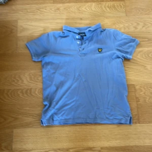Blå Lyle & Scott Junior pikétröja - Snygg ljusblå pikétröja från Lyle & Scott Junior med klassisk krage och tre knappar framtill. T-shirten har korta ärmar och den ikoniska gula örn-loggan broderad på bröstet. Tillverkad i mjuk bomull som är skön mot huden. Storlek 10/11 år men passar 152 också