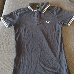 Mörkblå Fred Perry pike med krage - Snygg mörkblå pike från Fred Perry med klassisk krage och vita ränder på ärmslut och krage. Ikonisk Fred Perry-logga broderad på bröstet. Modellen har korta ärmar och är gjord i stretchig bomull för skön passform.