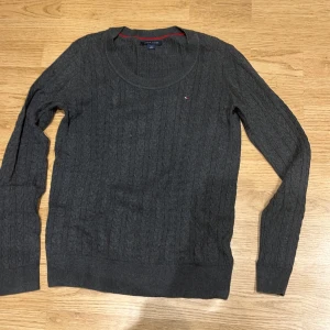 Grå kabelstickad tröja Tommy Hilfiger - Mörkgrå kabelstickad tröja från Tommy Hilfiger med rund halsringning och diskret logga på bröstet. Tröjan har långa ärmar och ribbade muddar. Perfekt för dig som gillar klassisk och stilren look.