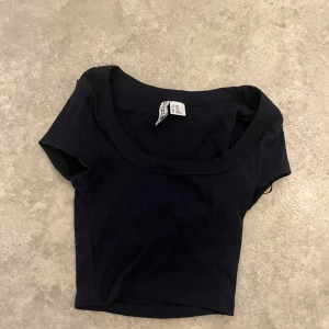 Svart croppad topp från Divided - En svart, croppad topp från Divided med korta ärmar och rundad halsringning. Toppen är ribbad och har en tight passform som sitter snyggt, dock är toppen väldigt kort. Perfekt att matcha med höga jeans eller kjol för en clean look.
