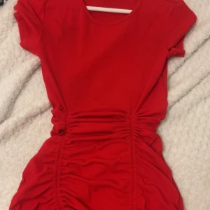 Röd kortklänning med snörning - Säljer en snygg röd kortklänning med rund hals och korta ärmar. Klänningen har coola rynkade detaljer och snörning längs sidorna som ger en trendig look. Materialet är mjukt och stretchigt, perfekt för dig som gillar att sticka ut med färg.