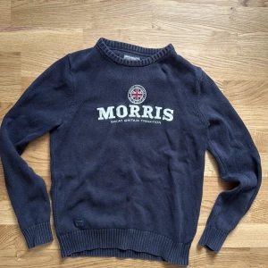 Mörkblå stickad tröja från Morris - Snygg mörkblå stickad tröja från Morris med rund hals och ribbade muddar. Framsidan har Morris-logga och ett broderat emblem med brittisk flagga. Perfekt för dig som gillar klassisk stil med en modern touch.